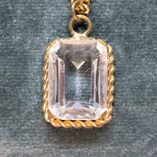 A close-up of a clear stone emerald cut pendant, within a twisted style gold-coloured setting, on a velvet background.