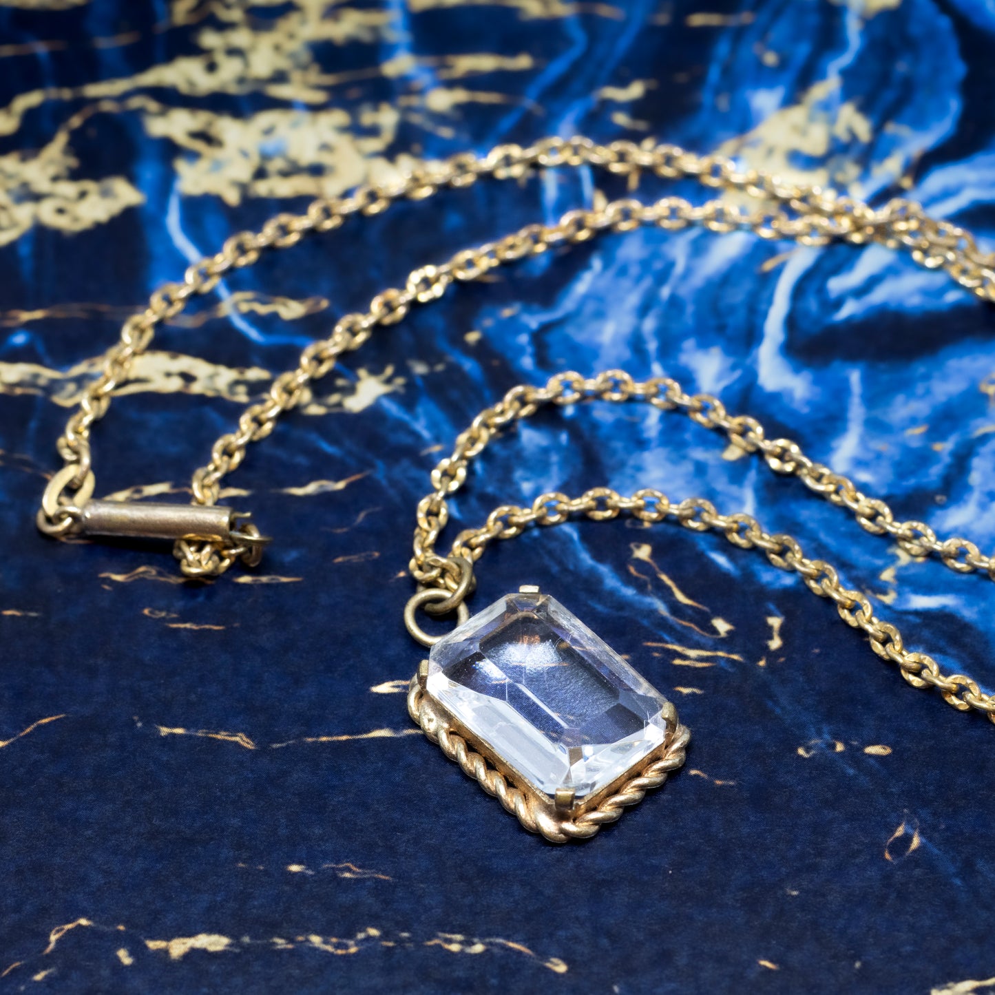 An emerald cut pendant on a gold-coloured chain, on a blue and gold marble background.