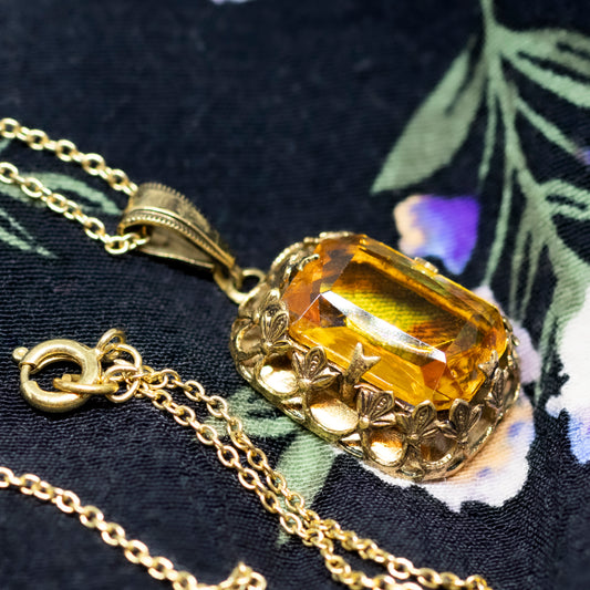 The side view of a radiant cut orange stone pendant, within a gold-coloured decorative frame, on a chain on a black and purple floral background.