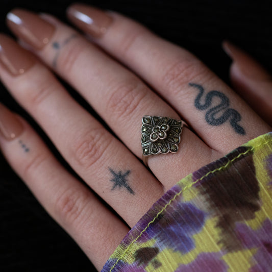 A silver tiered Art Deco style cocktail ring, set with marcasites, on the middle finger of a tattooed hand.