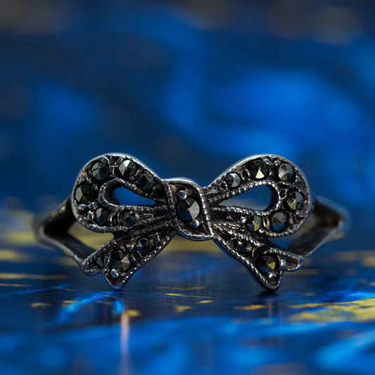 A silver bow design ring with split shoulders, set with marcasites, on a blue background.