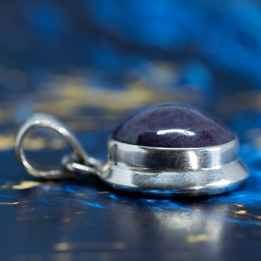 The side profile of a silver pendant, collet set with an oval cabochon amethyst, laid on a blue and gold marble surface.