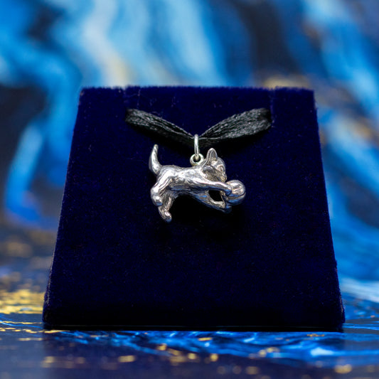 A silver cat pendant playing with a ball, hanging from a black ribbon, on a blue velvet stand.