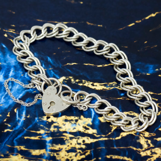 A silver double link curb bracelet with a heart padlock clasp and safety chain, laid on a blue and gold marble surface.