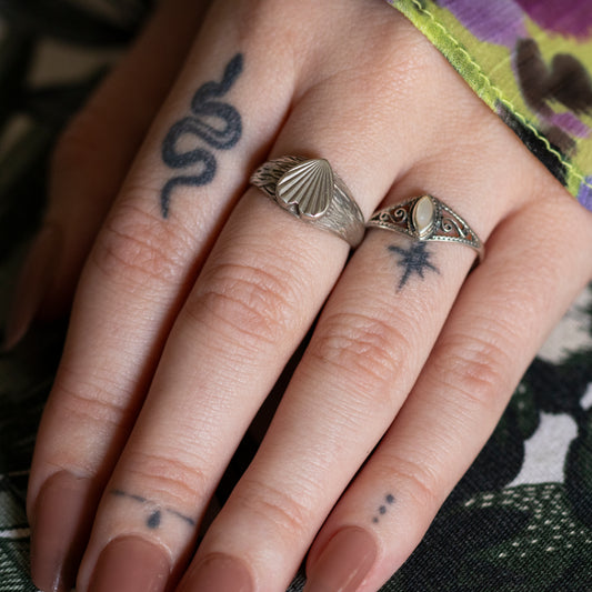A tattooed hand wearing a silver mother of pearl ring and a silver textured heart shape signet style ring.