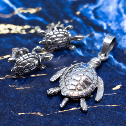 A silver turtle pendant with articulated limbs, together with a pair of matching stud earrings, on a blue and gold marble background.