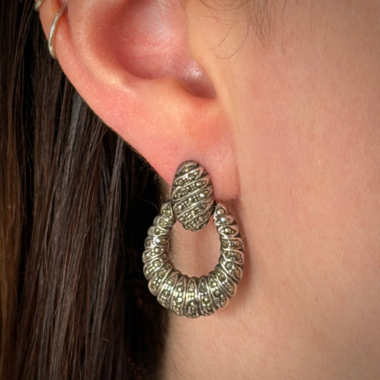 A close-up of a textured silver drop earring worn on a person's ear, showing the detailed spiral texture and sparkling marcasite accents.