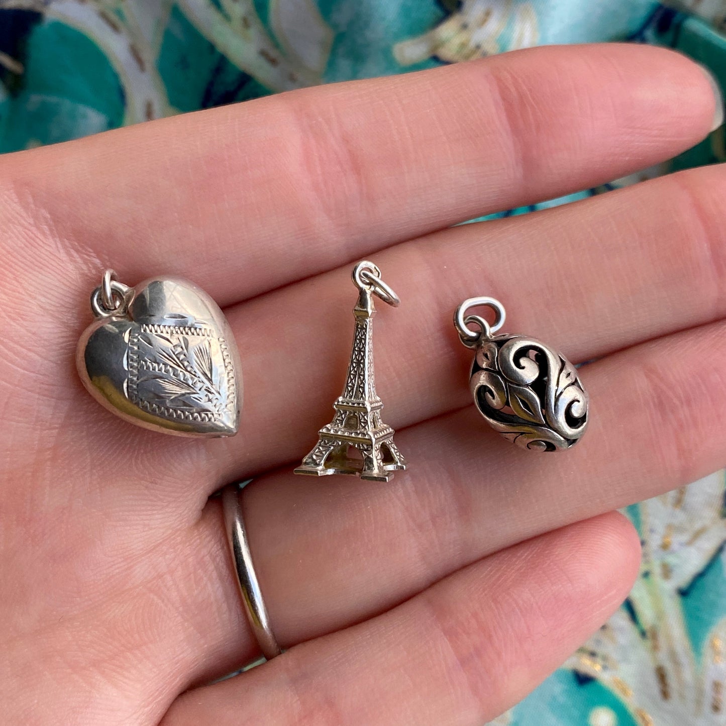 Three silver charms including a heart, Eiffel Tower, and spherical design on a hand.