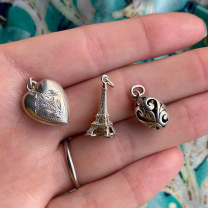 Three silver charms including a heart, Eiffel Tower, and spherical design on a hand.