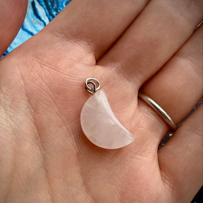 A small crescent moon pendant made of pale pink stone with a silver loop, resting on an open hand.