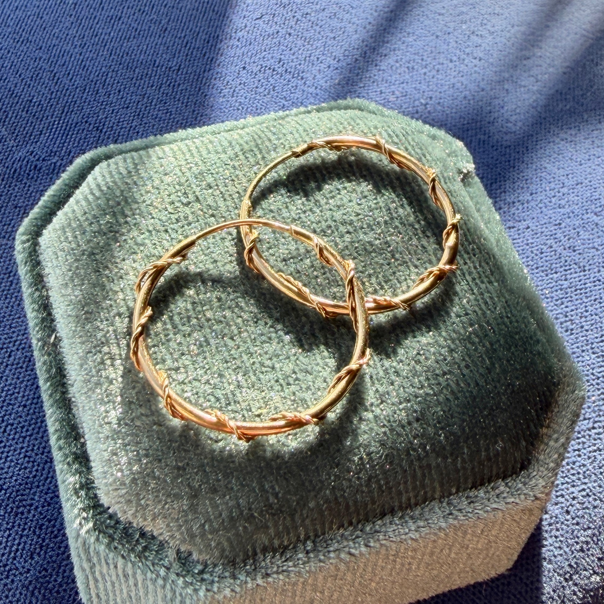 Rose gold hoop earrings on a green velvet cushion with a blue fabric background