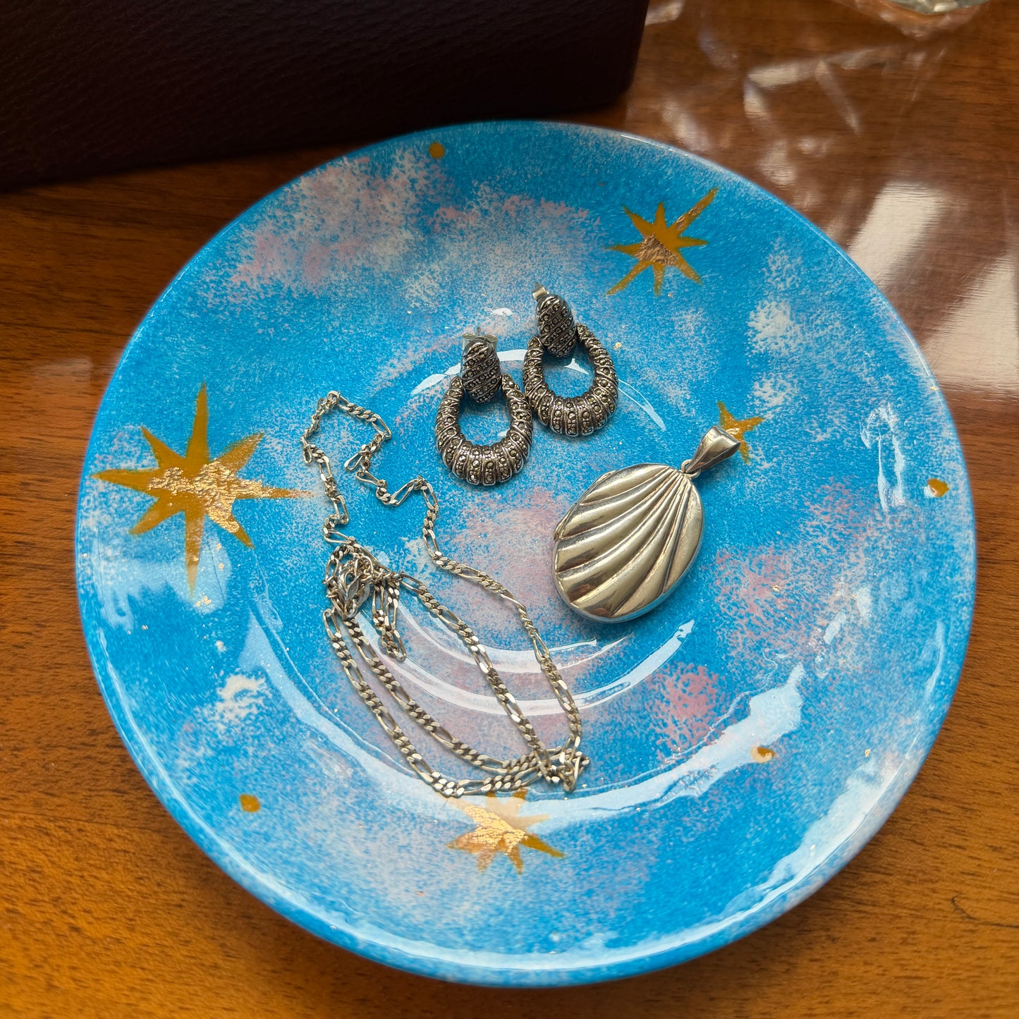 A jewelry set including a shell-shaped pendant necklace, textured hoop earrings, and a chain necklace, displayed on a blue ceramic plate with gold star details, set on a wooden table.
