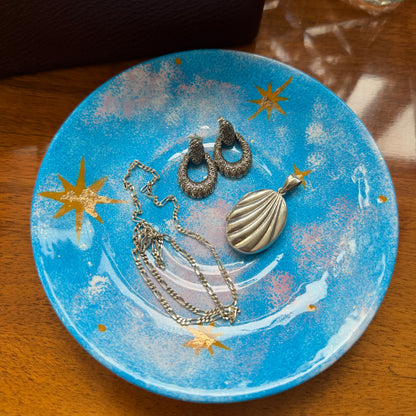 A jewelry set including a shell-shaped pendant necklace, textured hoop earrings, and a chain necklace, displayed on a blue ceramic plate with gold star details, set on a wooden table.
