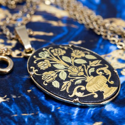 The side view of an oval black and gold-coloured damascene pendant on a blue background.
