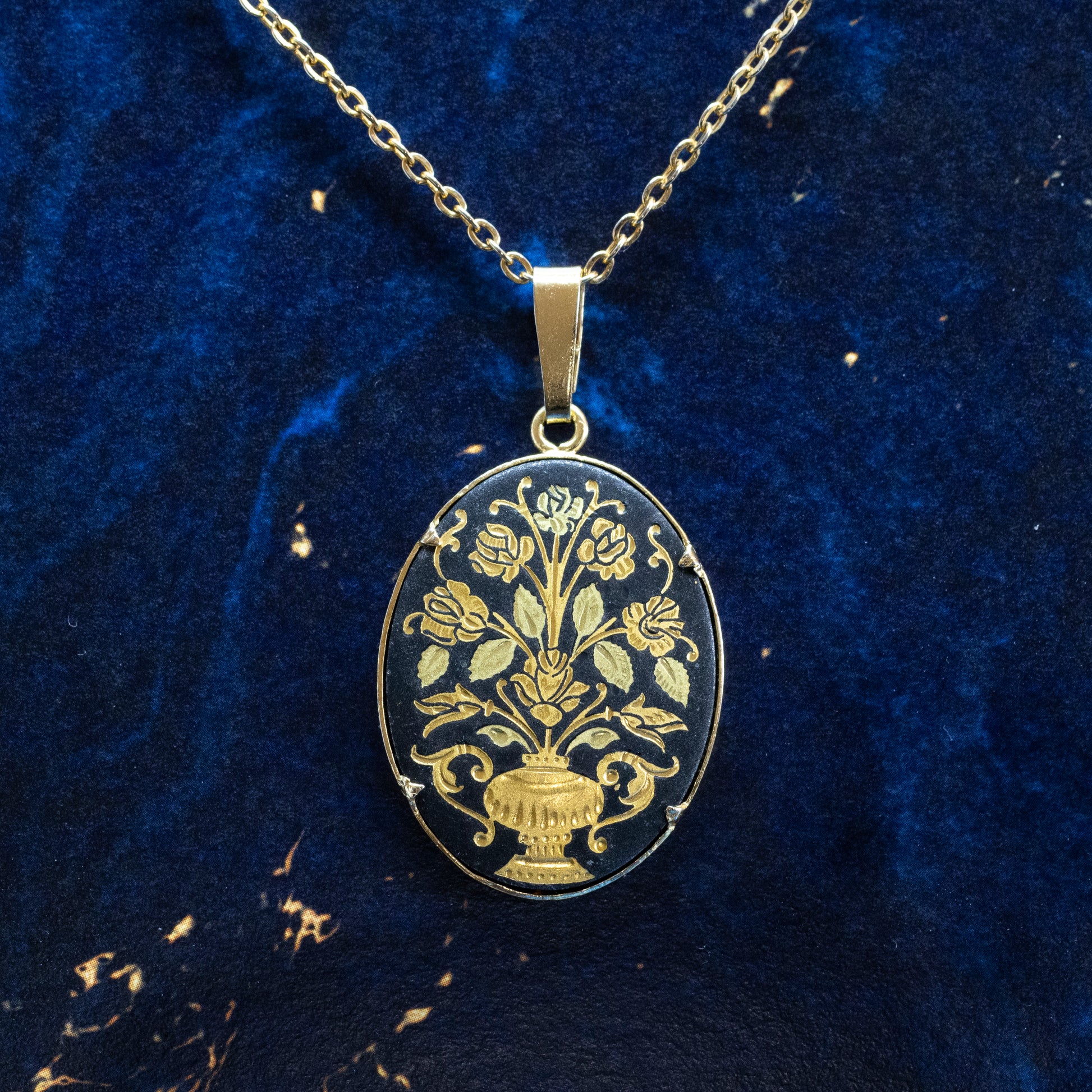 An oval black and gold-coloured damascene pendant, hanging from a chain on a blue background.