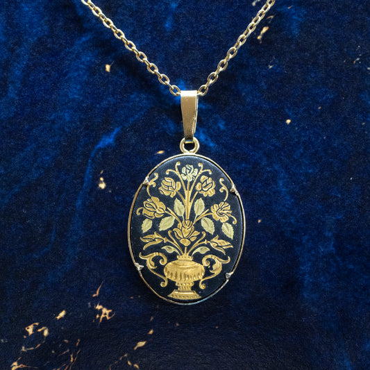 An oval black and gold-coloured damascene pendant, hanging from a chain on a blue background.