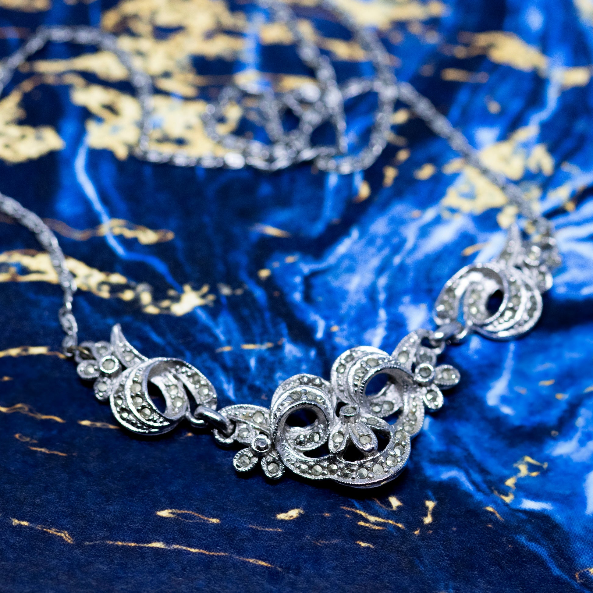 A vintage style silver and marcasite collar necklace, on a blue and gold marble background.