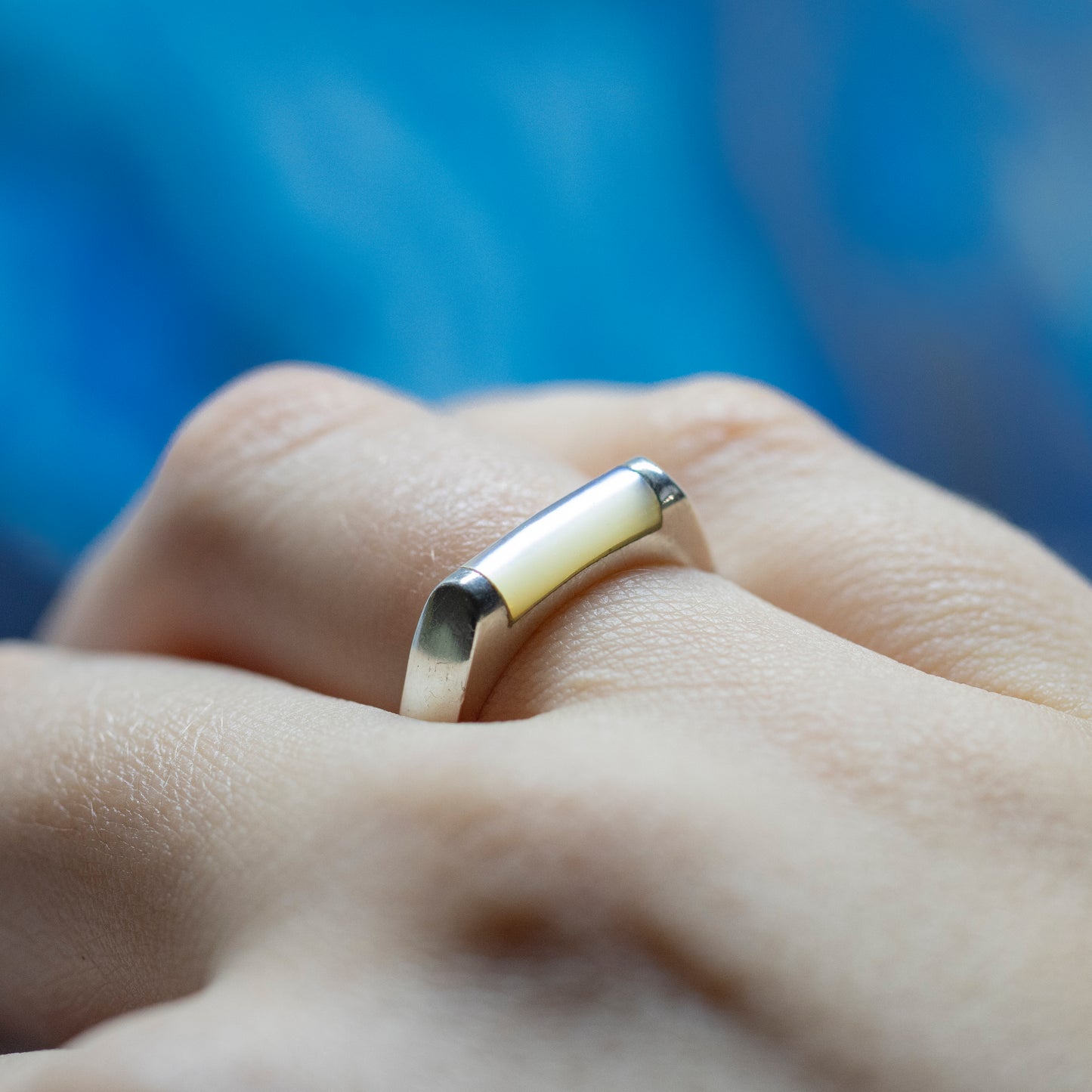 A close-up of a silver mother of pearl bar ring from the side, being worn on a hand with a blue background.