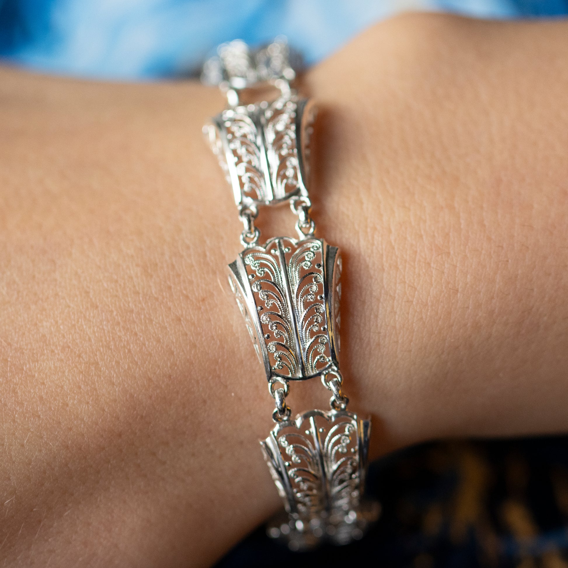 A wrist wearing an intricate filigree design silver panel bracelet, with a blue background.