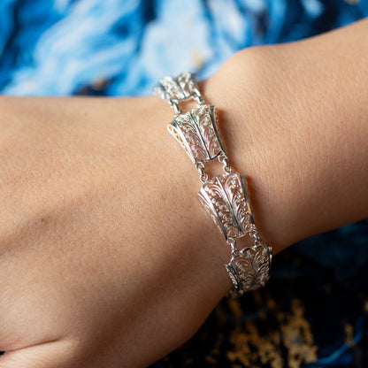 A wrist wearing an intricate filigree design silver panel bracelet, with a blue background.