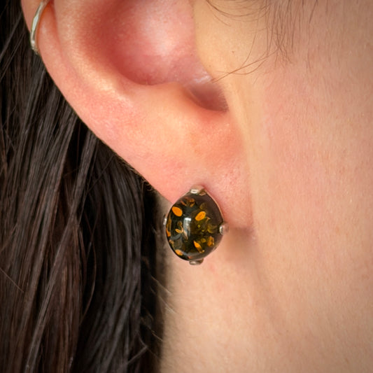 A close-up of a person's ear wearing a small oval-shaped earring with a dark amber-brown center and scattered gold flecks, set in a silver-toned metal.