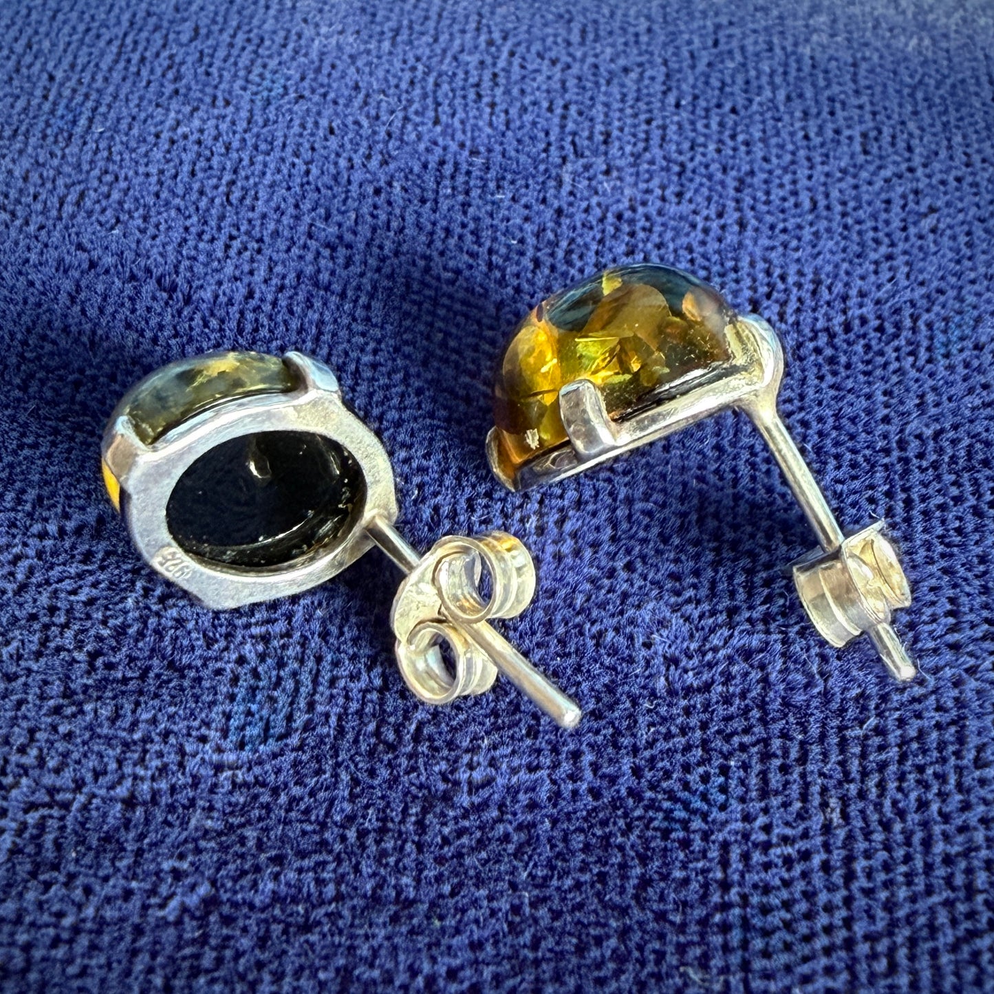 A pair of silver stud earrings with oval-cut amber gemstones, shown on a textured blue fabric background.