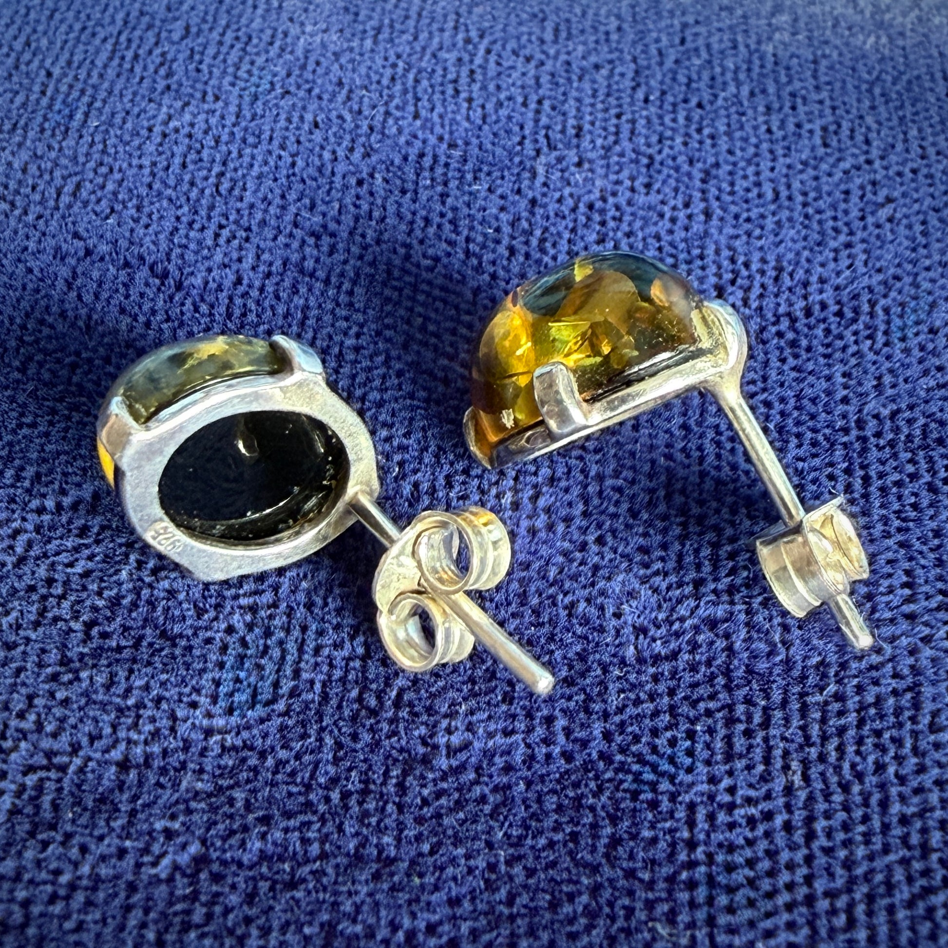 A pair of silver stud earrings with oval-cut amber gemstones, shown on a textured blue fabric background.