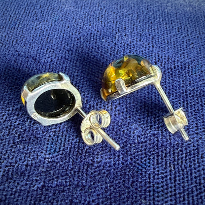 A pair of silver stud earrings with oval-cut amber gemstones, shown on a textured blue fabric background.