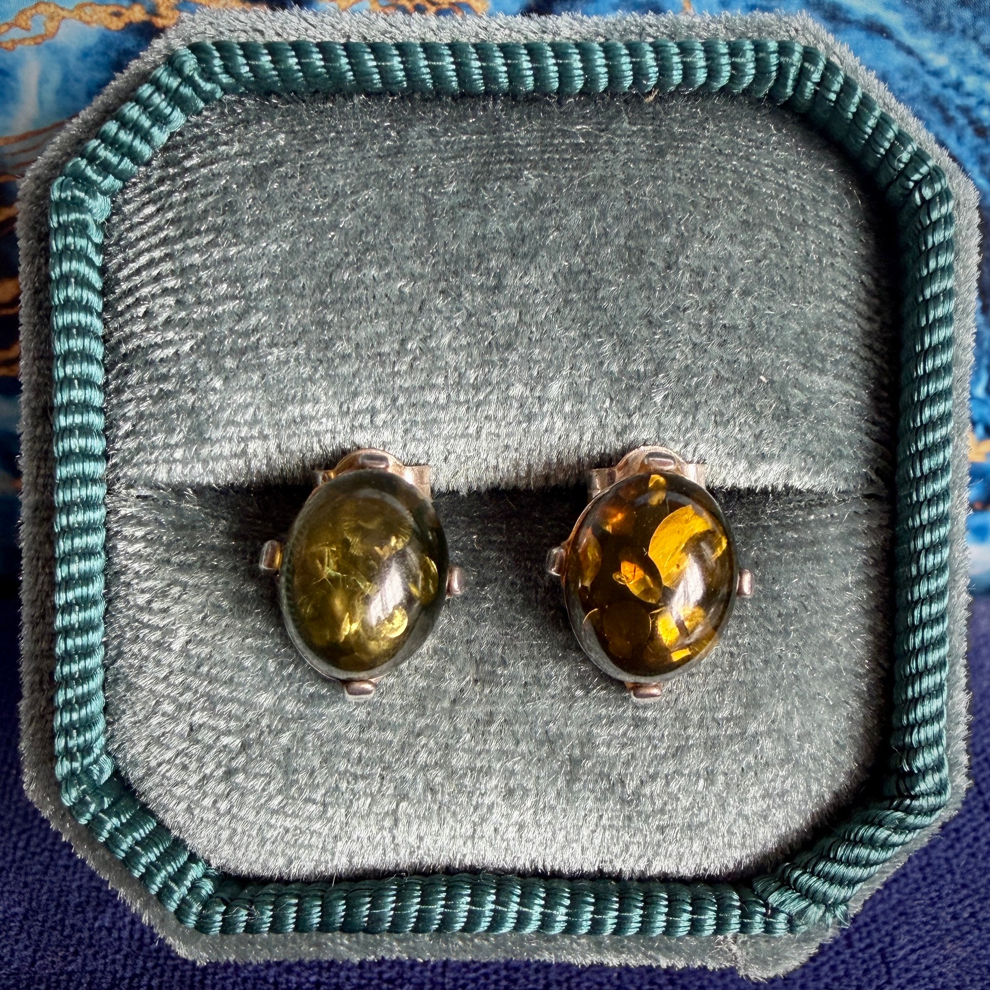 A pair of oval stud earrings with amber-colored stones in silver settings, displayed on a gray velvet jewelry box with a teal fabric border.