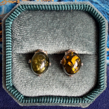 A pair of oval stud earrings with amber-colored stones in silver settings, displayed on a gray velvet jewelry box with a teal fabric border.