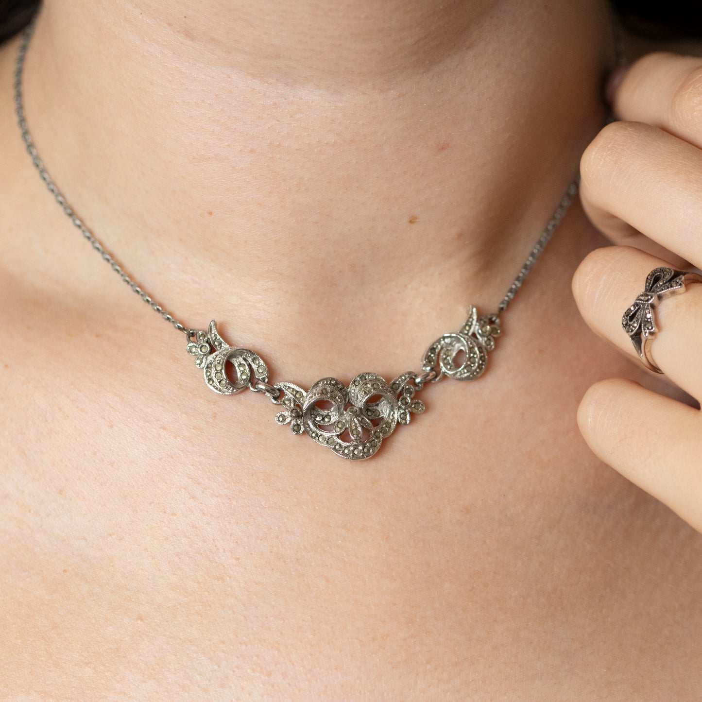 A vintage style silver and marcasite collar necklace, being worn on a neck, with a hand wearing a silver and marcasite bow design ring.
