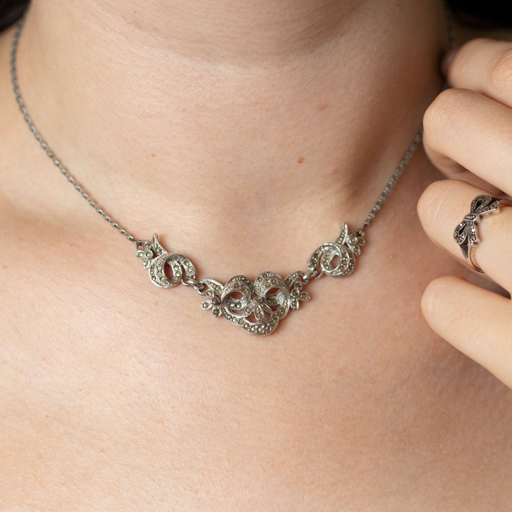A vintage style silver and marcasite collar necklace, being worn on a neck, with a hand wearing a silver and marcasite bow design ring.