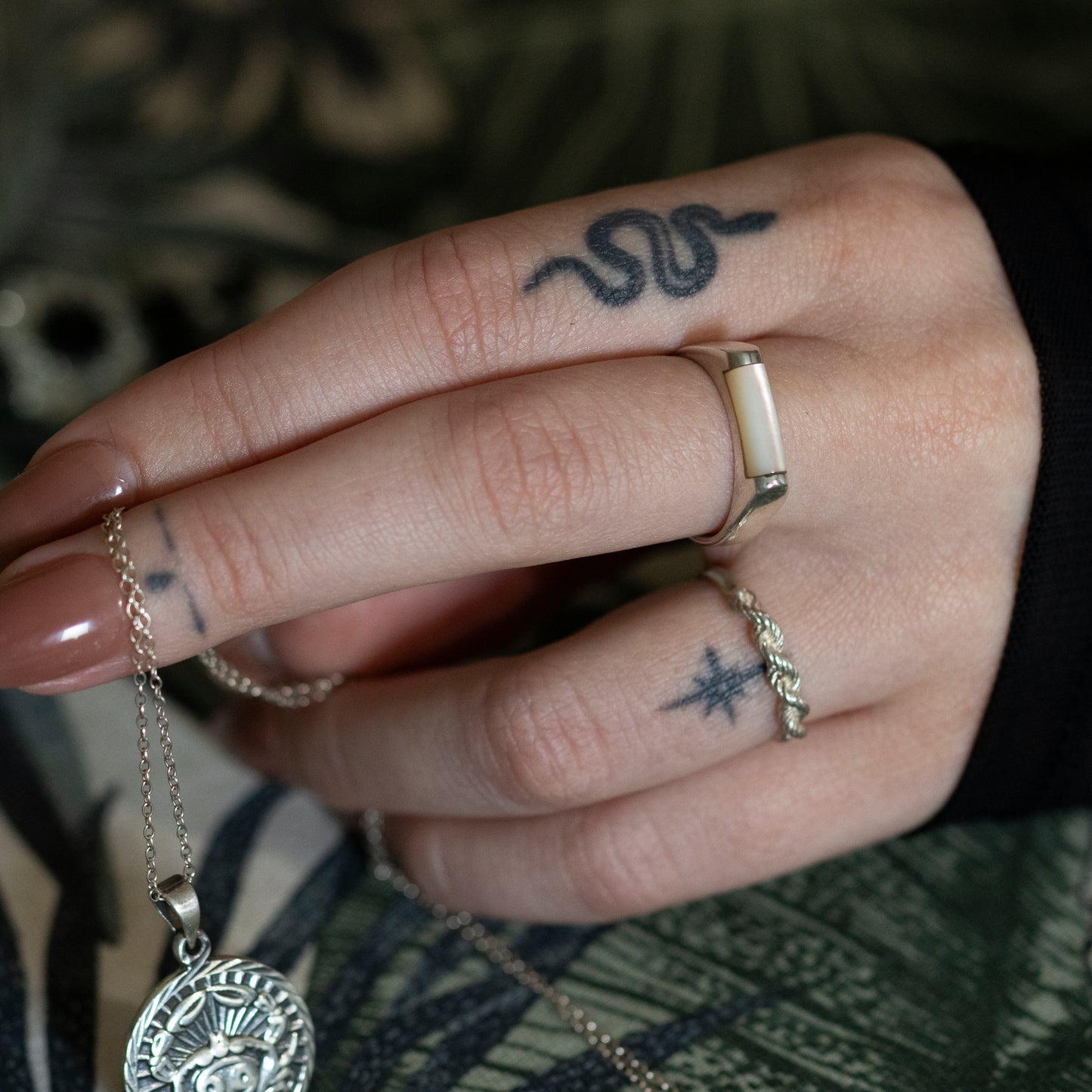 A tattooed hand wearing a silver mother of pearl bar ring and a silver twisted rope design ring, holding a silver medallion style necklace.