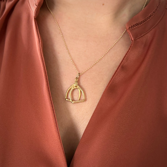 A woman wearing a gold stirrups pendant on a fine gold chain and a peach silk shirt.