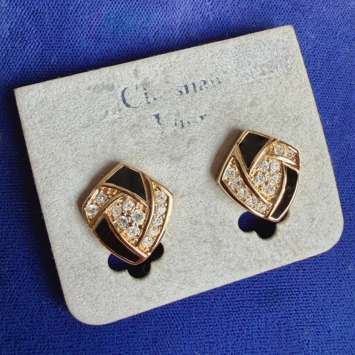 A pair of vintage shield-shaped gold-tone earrings with black enamel and clear crystal accents, displayed on a light gray jewelry card against a textured blue background.