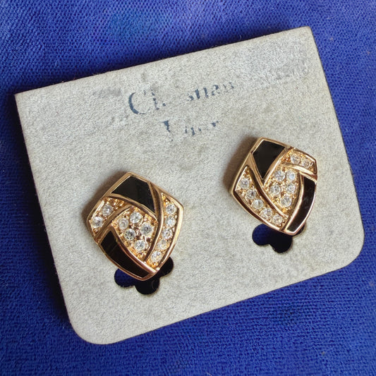 A pair of vintage shield-shaped gold-tone earrings with black enamel and clear crystal accents, displayed on a light gray jewelry card against a textured blue background.